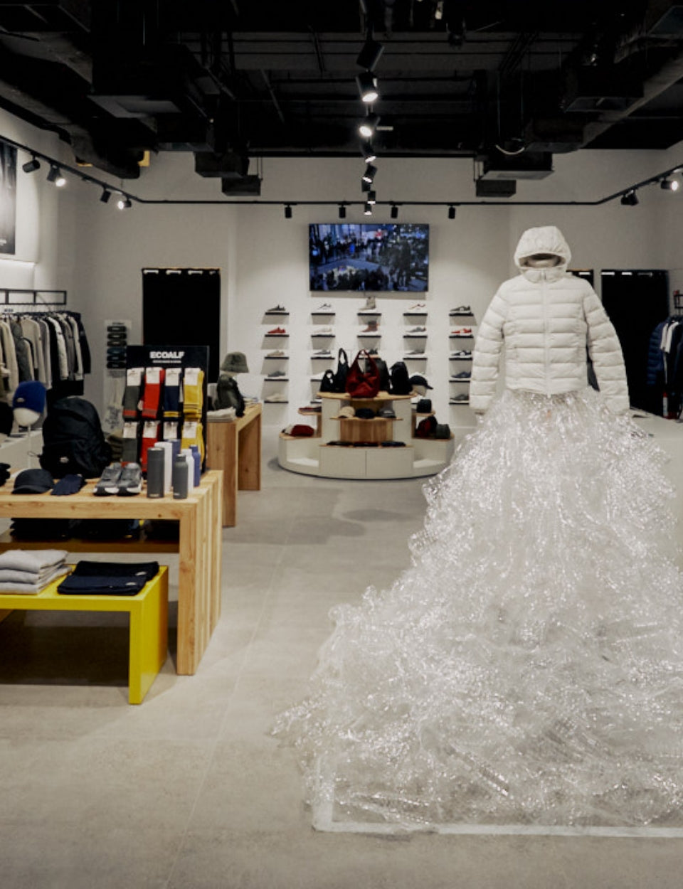 Interior de una tienda Ecoalf con un abrigo blanco con capucha sobre un montaje de plástico reciclado; al fondo, estantes de zapatos y accesorios y una pantalla con imágenes.