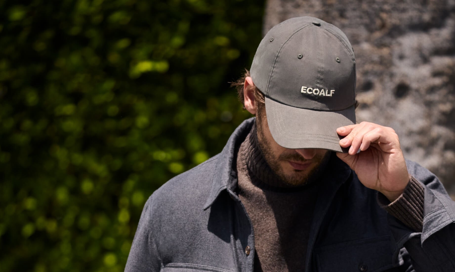 Un hombre con gorra gris, chaqueta técnica y jersey marrón sostenible de la marca Ecoalf en un entorno natural.