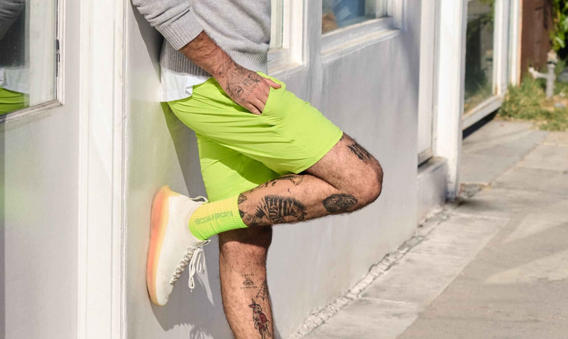 Un hombre vistiendo ropa deportiva sostenible de la marca Ecoalf, con zapatillas y calcetines verde flúor junto a un edificio moderno.