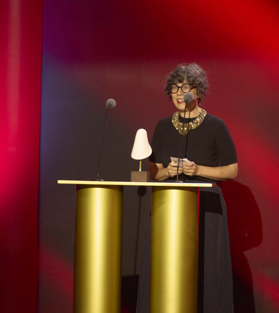 Una mujer con gafas negras y un vestido negro con collar dorado habla tras un atril dorado con dos micrófonos, en un escenario iluminado de rojo.