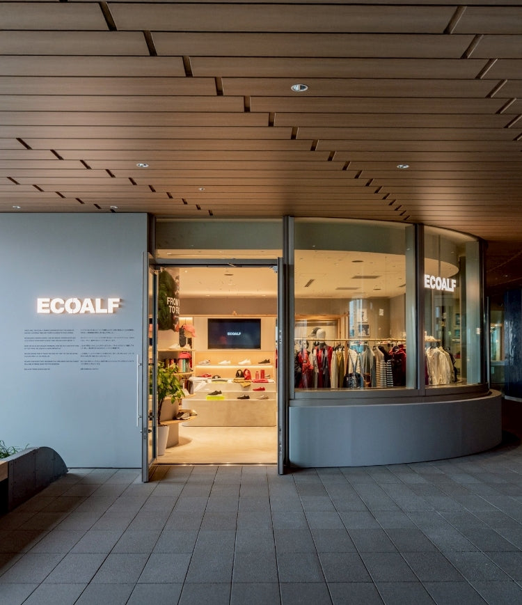Entrada de una tienda Ecoalf moderna con techo de madera; por las puertas de vidrio se ve el interior iluminado con ropa y zapatos, y el logo "ECOALF" en la pared.