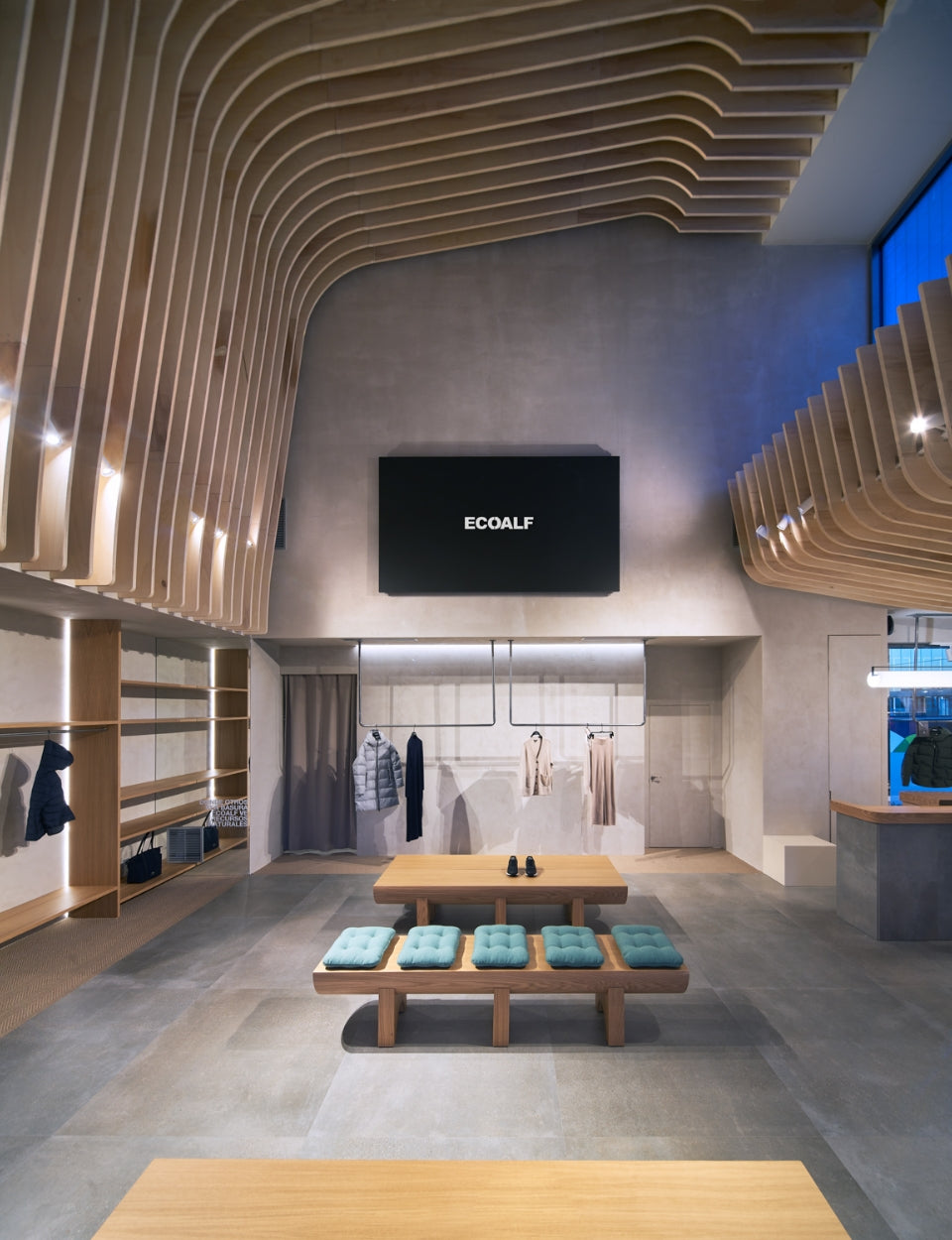Una tienda Ecoalf moderna y minimalista con techo de listones de madera curvados, un banco de madera con cojines verde azulado y ropa colgada al fondo.