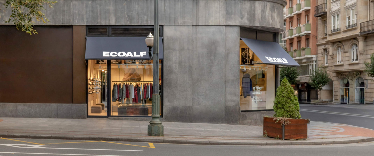 Fachada en esquina de una tienda Ecoalf con dos toldos negros y el rótulo "ECOALF"; por las ventanas se ve ropa colgada y, delante, un pequeño árbol en un macetero.