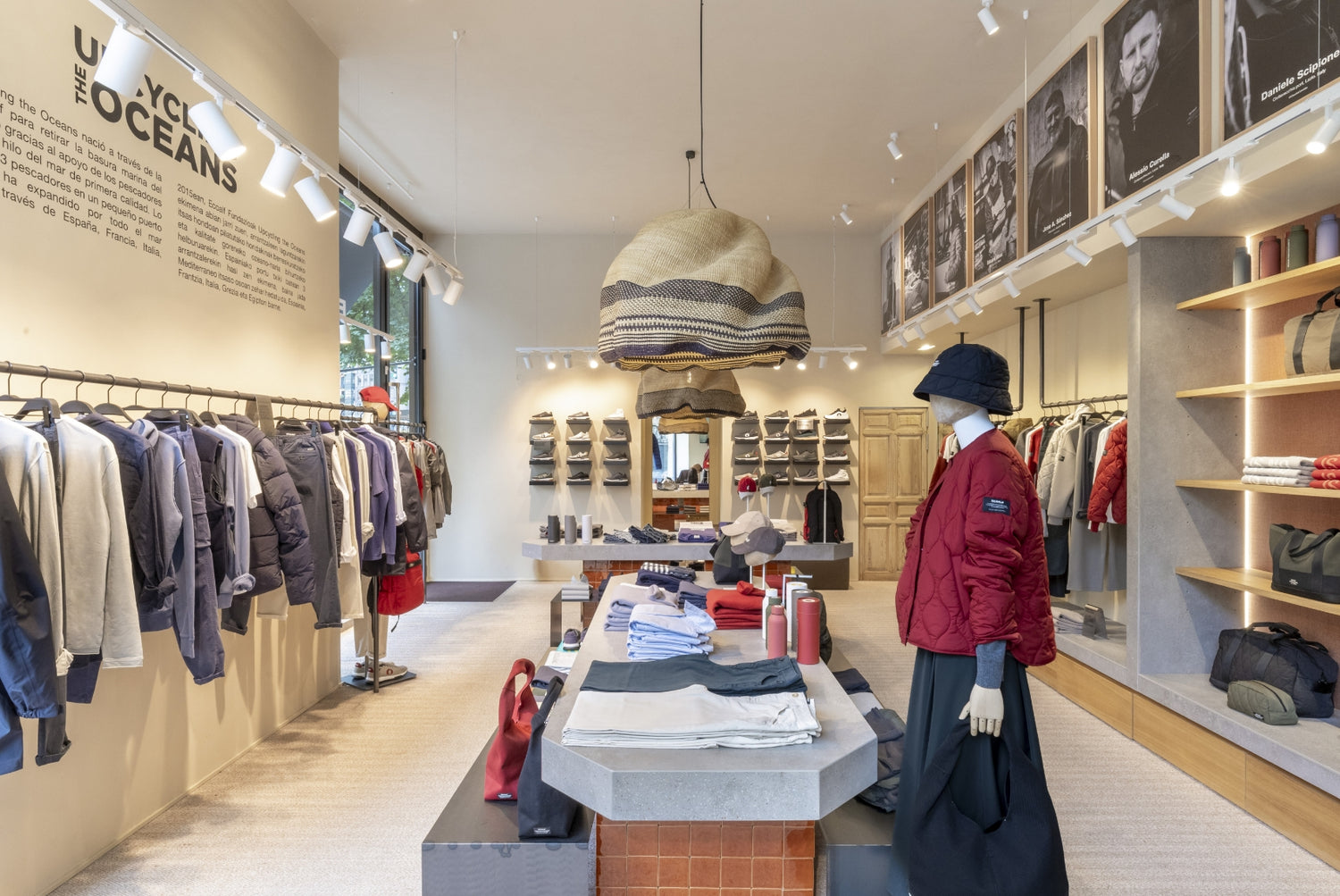 Interior luminoso de una tienda Ecoalf con chaquetas y sudaderas en las paredes, un maniquí con chaqueta roja, mesas con ropa doblada y botellas, y zapatos al fondo.