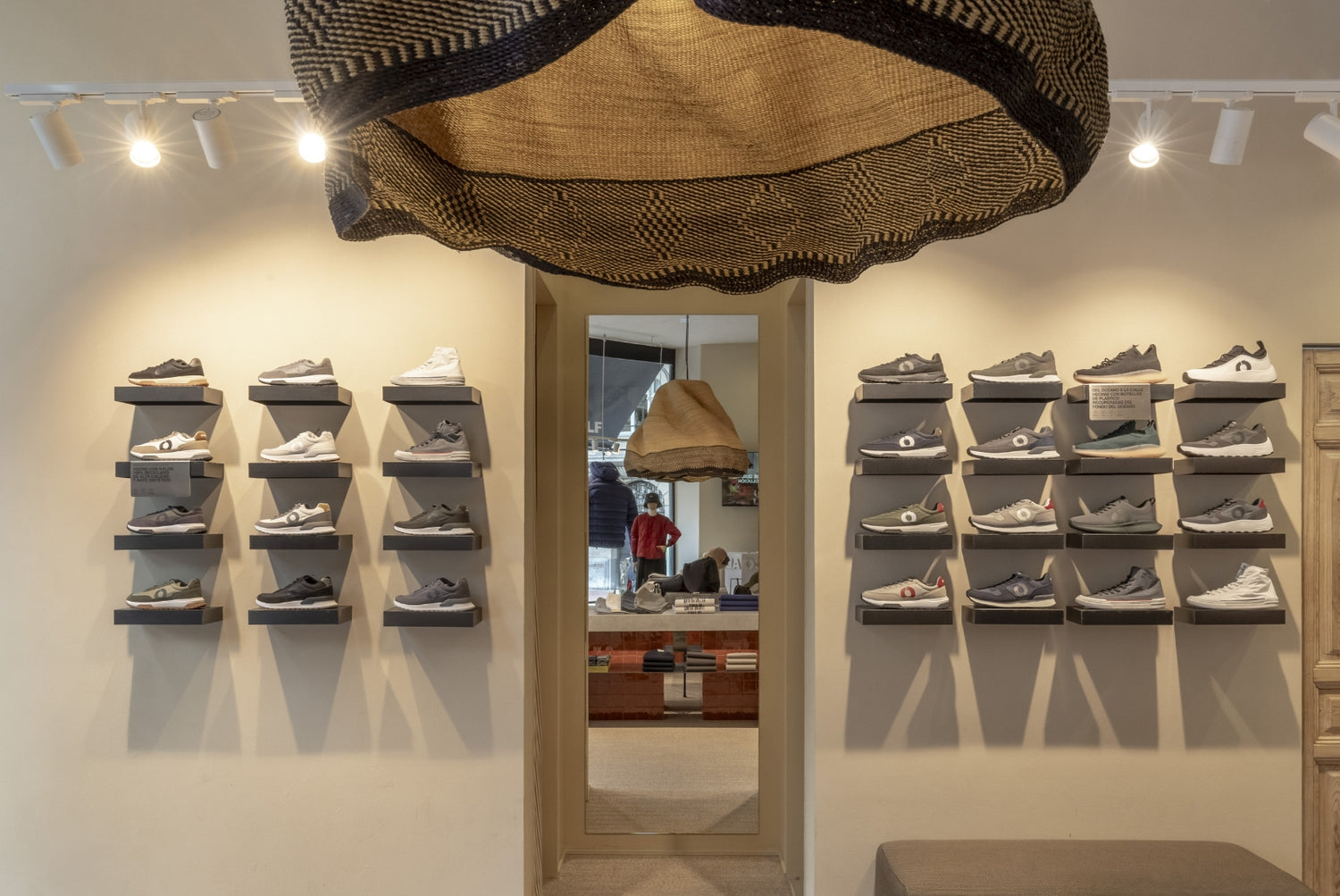 Una pared de tienda con zapatillas en tonos neutros expuestas en baldas individuales; encima, una lámpara tejida, y al fondo un gran espejo refleja el interior.