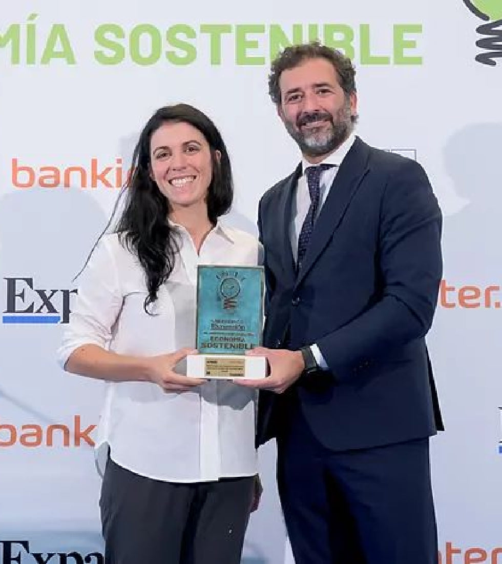 Una mujer con blusa blanca sostiene un trofeo con la placa "Economía Sostenible" junto a un hombre de traje oscuro; al fondo, logos corporativos y texto promocional.