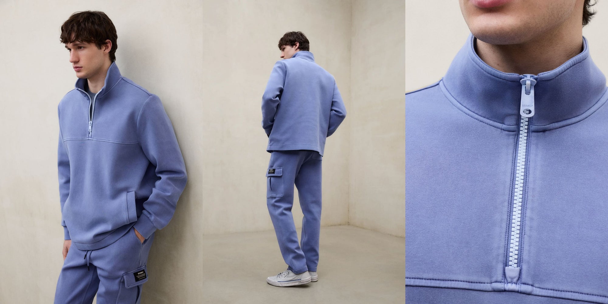Un hombre con un conjunto de Ecoalf azul de sudadera de cremallera y pantalón con etiqueta en el bolsillo y zapatillas blancas, en una sala de pared clara.