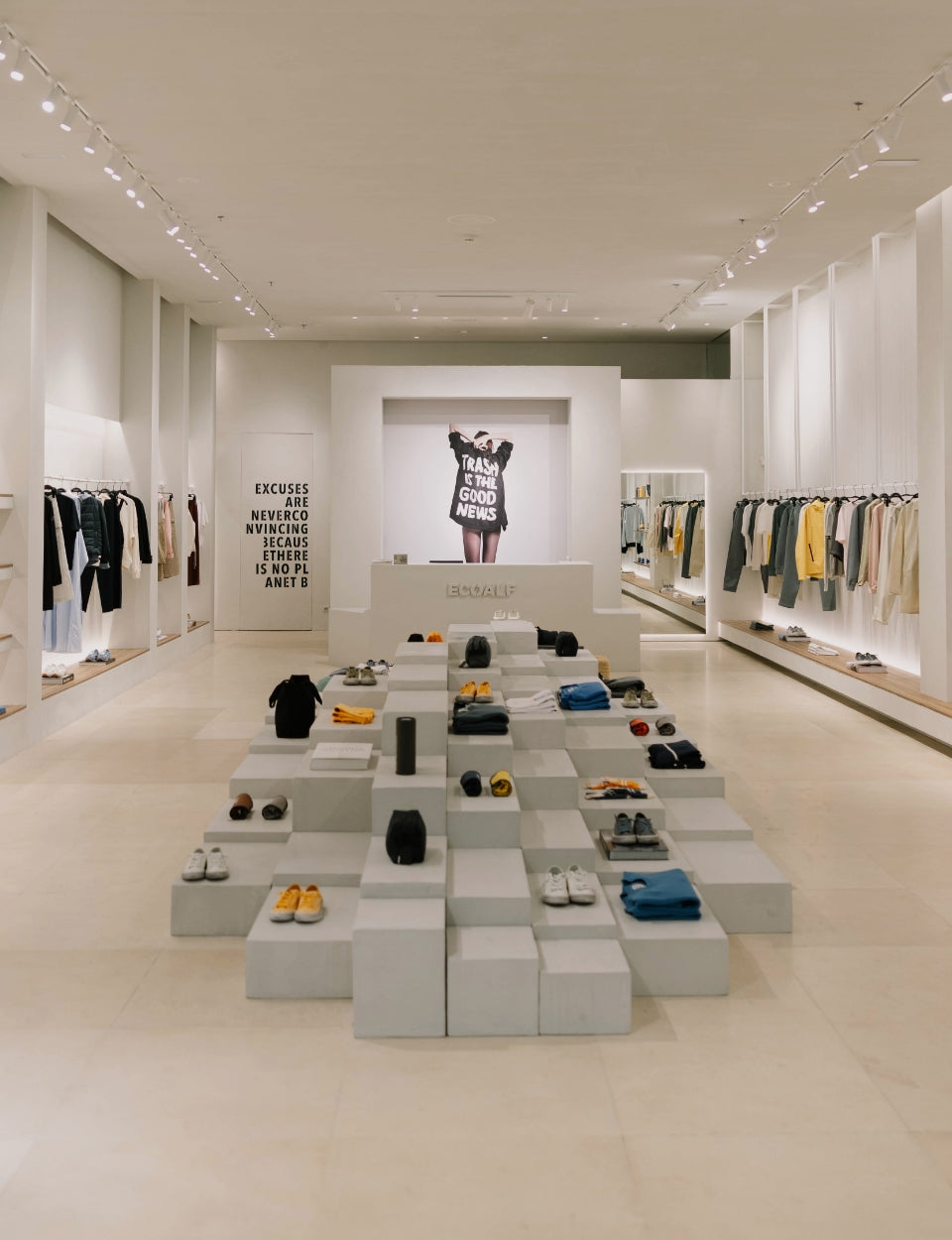 Una tienda Ecoalf moderna y minimalista de paredes blancas, con ropa y zapatos sobre bloques y estantes y, al fondo, un cartel con un mensaje motivacional.