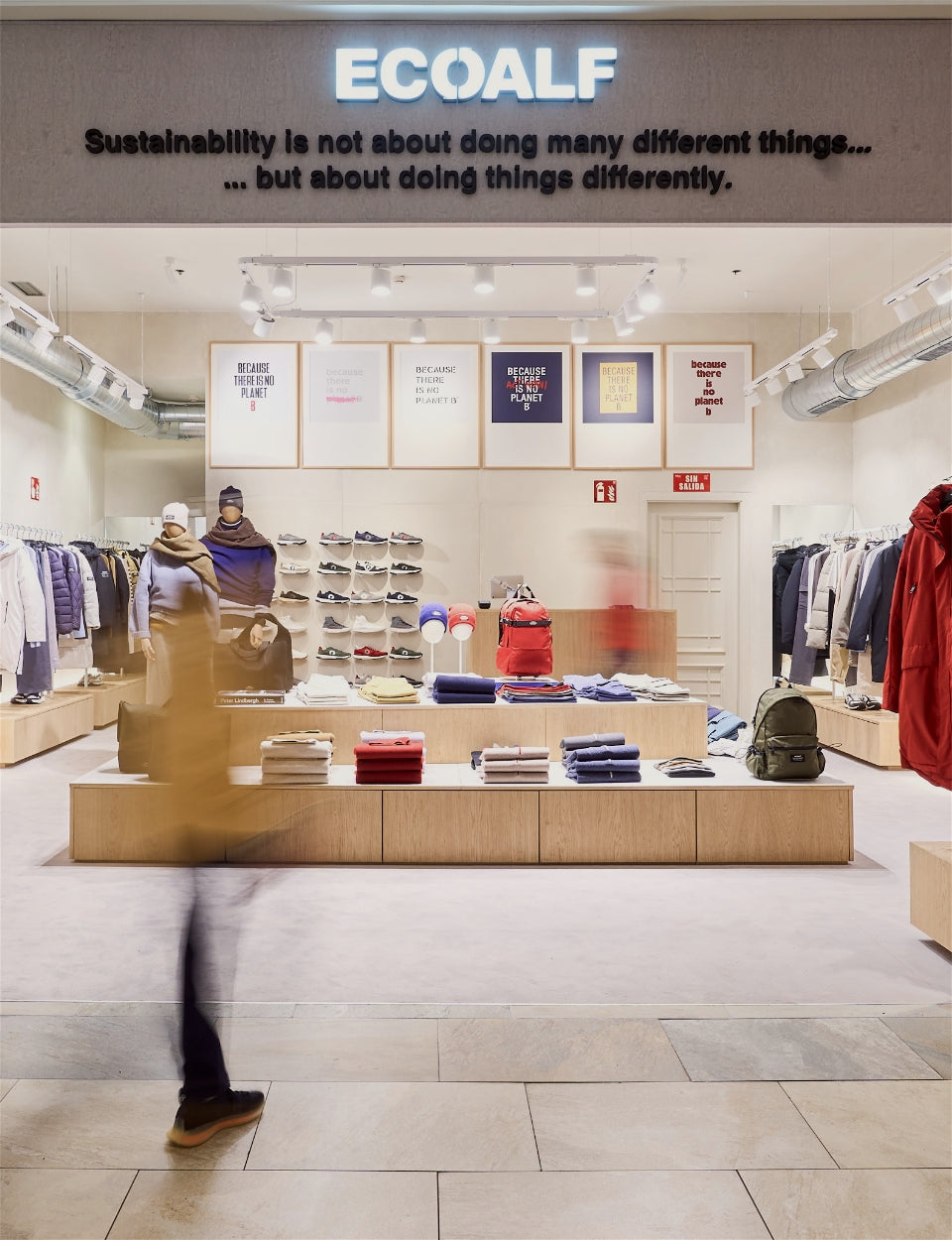 Una tienda Ecoalf moderna y ordenada, con frases sobre sostenibilidad en las paredes y prendas, mochilas y sombreros expuestos en estantes y mesas, con una persona al frente.