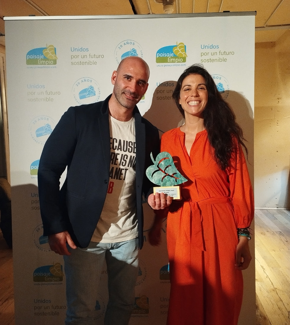 Una mujer con vestido rojo sostiene un trofeo con forma de hoja junto a un hombre con camiseta blanca y chaqueta oscura, ante un fondo promocional.
