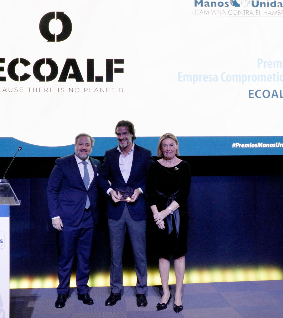 Tres personas en un escenario durante una entrega de premios; el hombre del centro sostiene un trofeo. Al fondo, una pantalla con el logo "ECOALF" y el texto del premio.