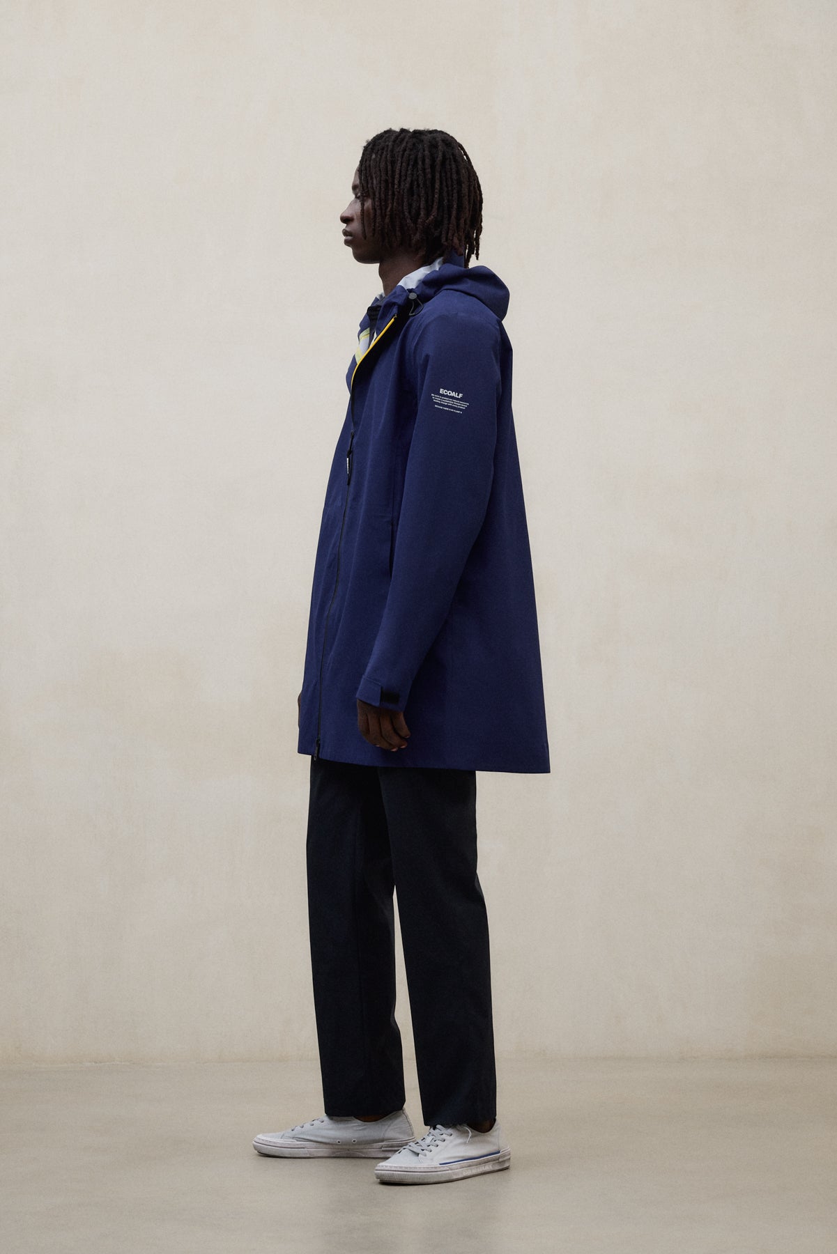 Un hombre de perfil con una chaqueta de Ecoalf azul con capucha, pantalón oscuro y zapatillas blancas, sobre un suelo claro y fondo beige.