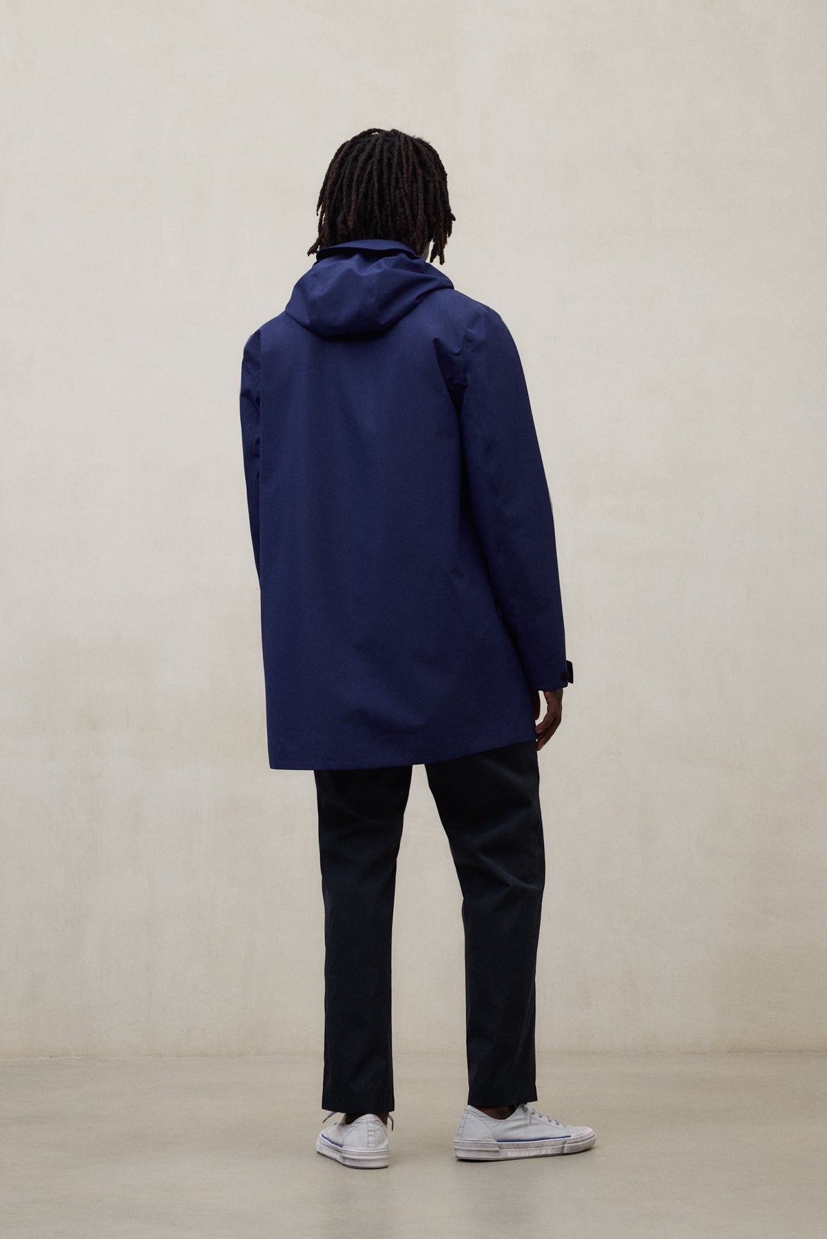 Un hombre de espaldas con una chaqueta de Ecoalf azul con capucha y pantalón oscuro y zapatillas blancas, en un entorno minimalista beige.