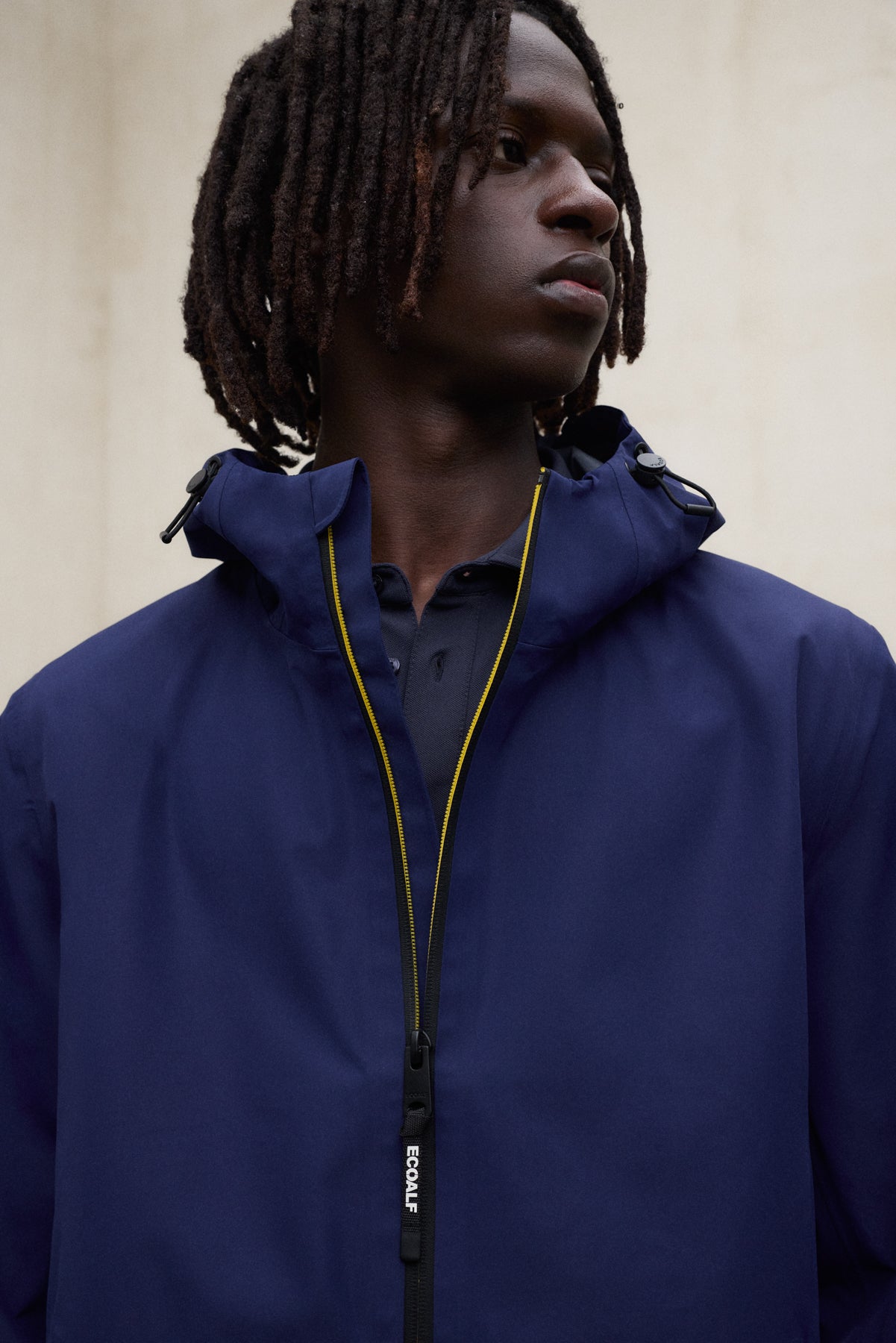 Un hombre de perfil con una chaqueta de Ecoalf azul oscuro con capucha y detalles de cremallera amarillos, sobre fondo neutro.