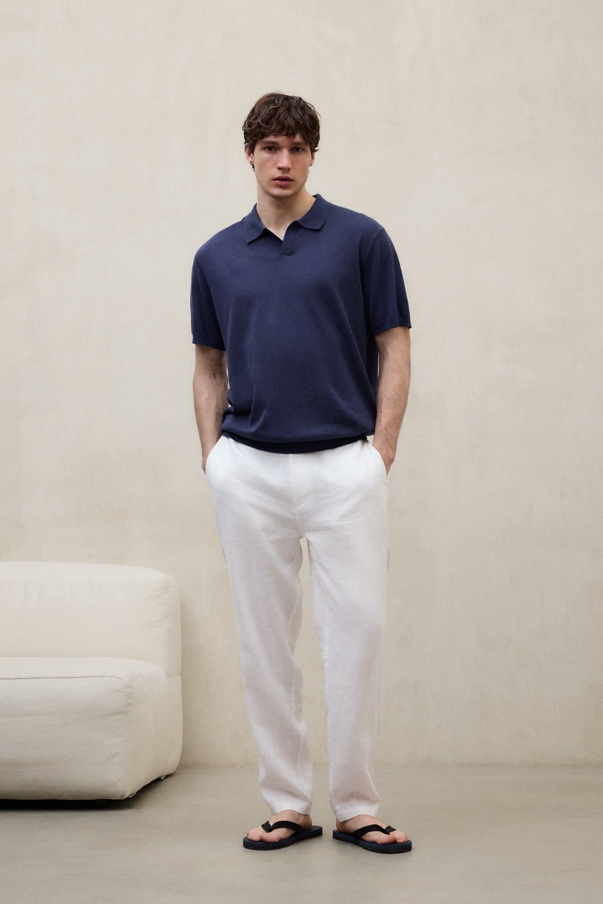 Un hombre con un polo de Ecoalf azul marino, pantalón blanco y sandalias negras, en un entorno minimalista con un sofá blanco al lado.