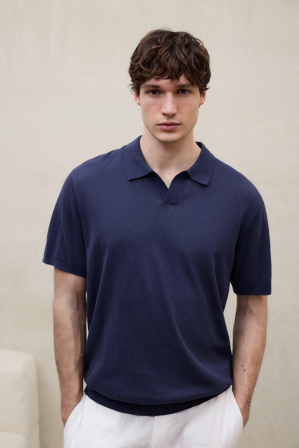 Un hombre con un polo de Ecoalf azul oscuro y las manos en los bolsillos del pantalón blanco, sobre fondo beige claro.