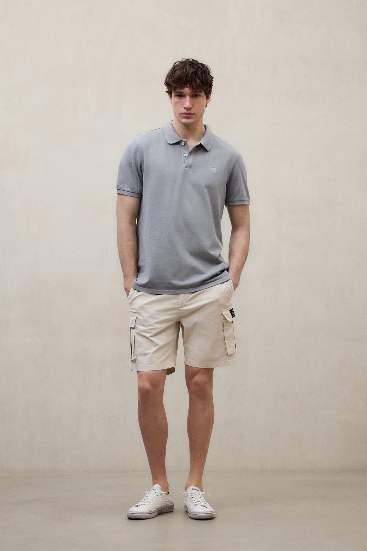 Un hombre con un polo de Ecoalf gris claro y shorts beige, con zapatillas blancas y las manos en los bolsillos, sobre un muro neutro.