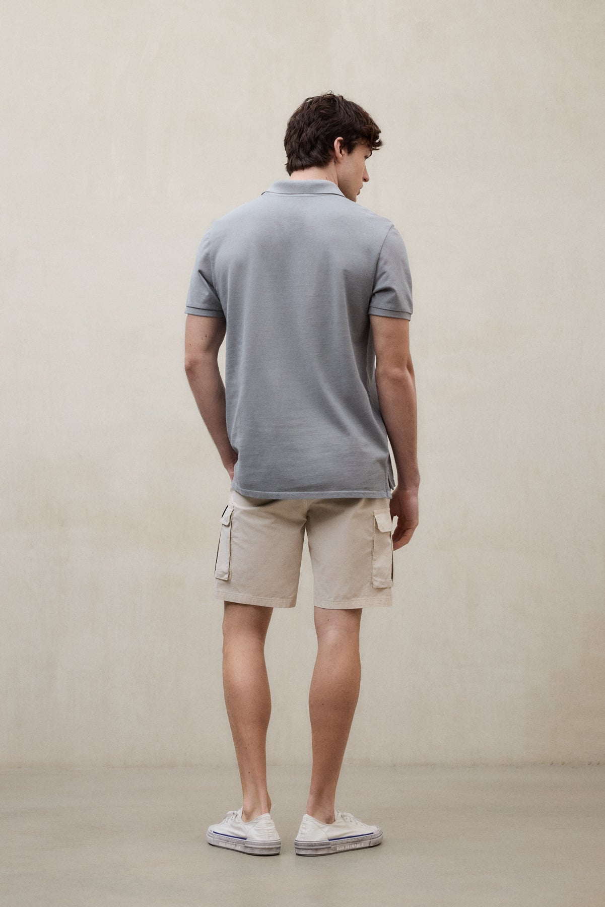 Un hombre de espaldas con un polo de Ecoalf gris y shorts beige con bolsillos laterales y zapatillas blancas, en una sala de paredes neutras.