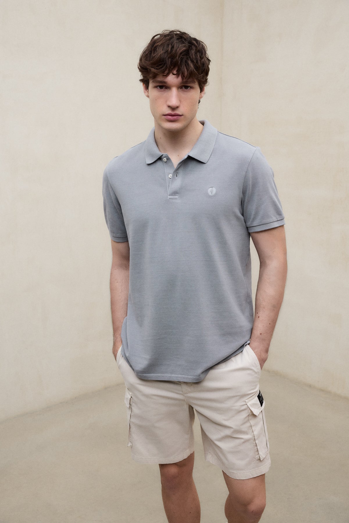 Un hombre con un polo de Ecoalf gris claro y shorts beige con bolsillos laterales, las manos en los bolsillos, sobre fondo neutro.