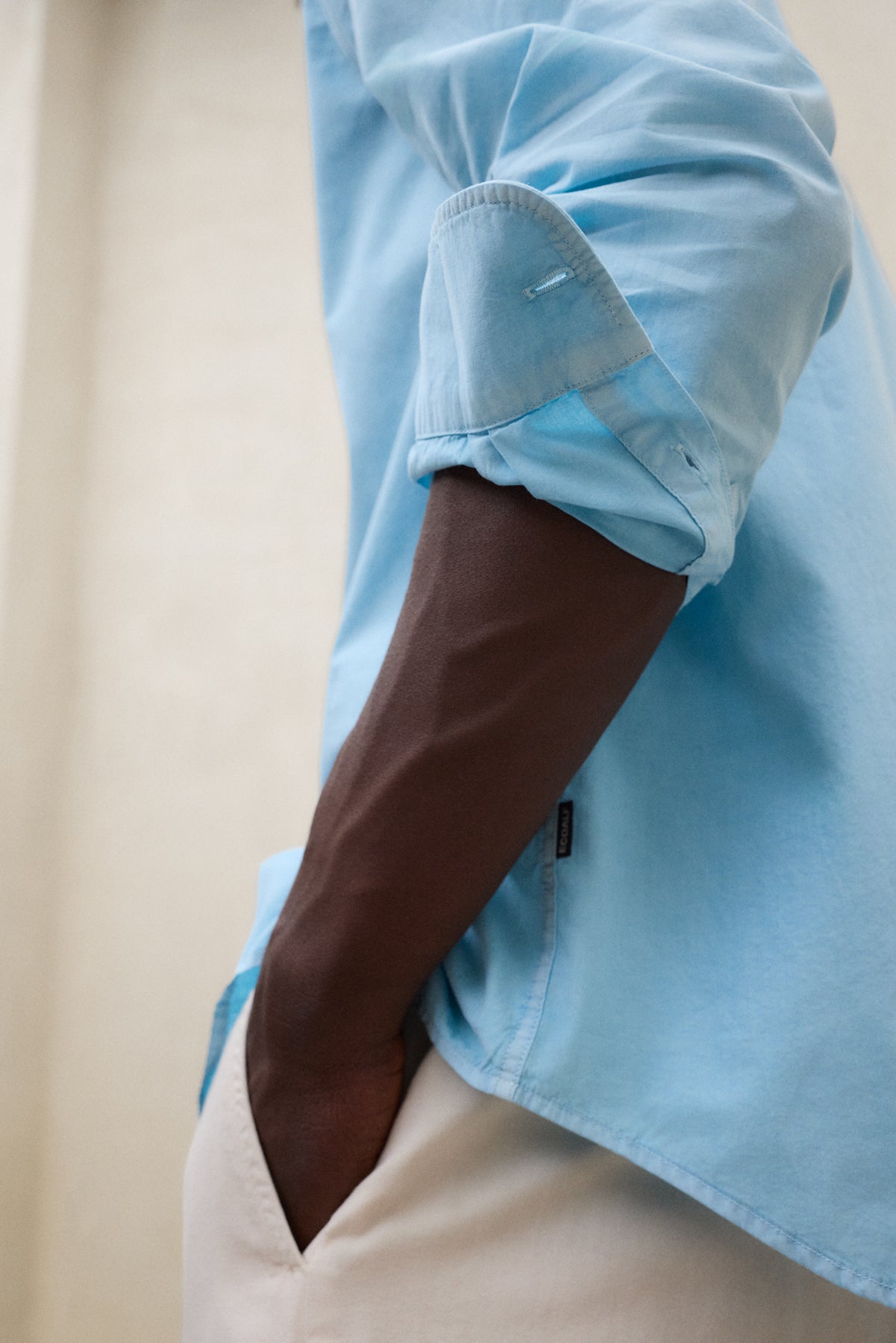 Camisa azul remangada de la firma Ecoalf y pantalón beige con la etiqueta de compromiso de la marca a la vista.