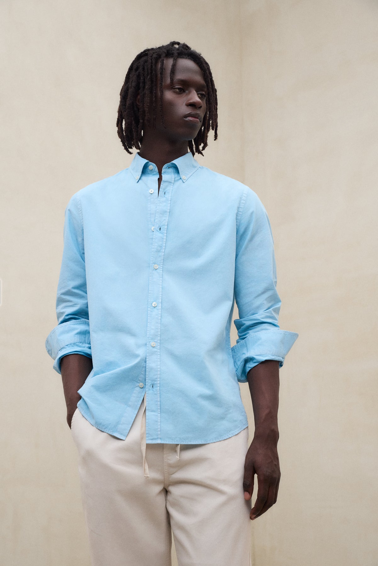 Un hombre con camisa de Ecoalf azul claro de manga larga y pantalón beige, sobre fondo neutro.