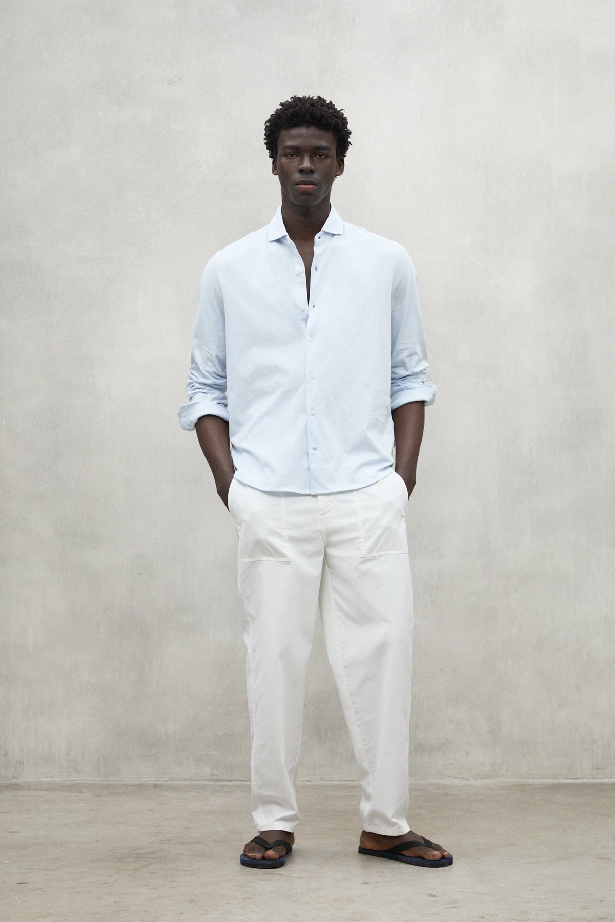 Un hombre posando con camisa azul claro de la marca Ecoalf y pantalón blanco de fibras recicladas sobre fondo gris.