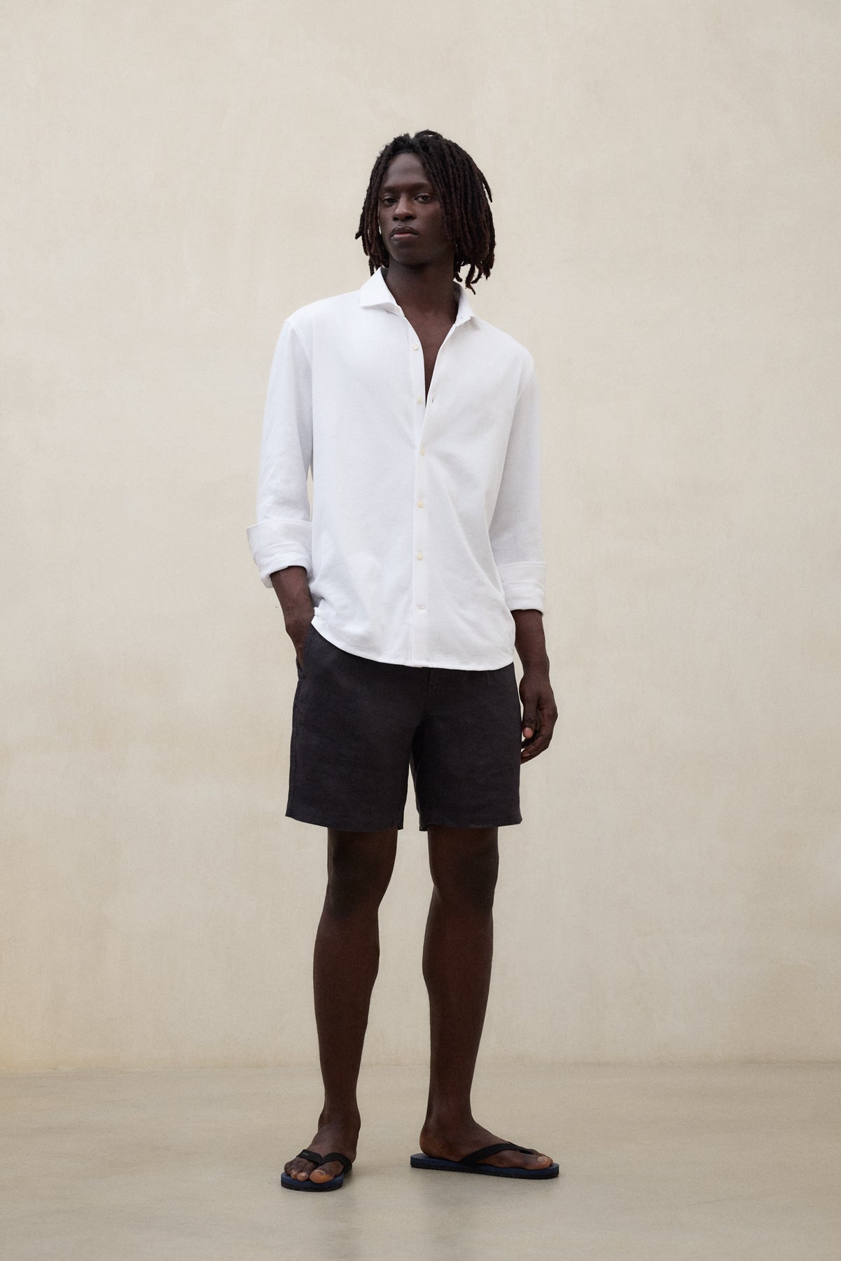 Un hombre con camisa de Ecoalf blanca de manga larga desabrochada, shorts oscuros y sandalias negras, sobre fondo beige liso.