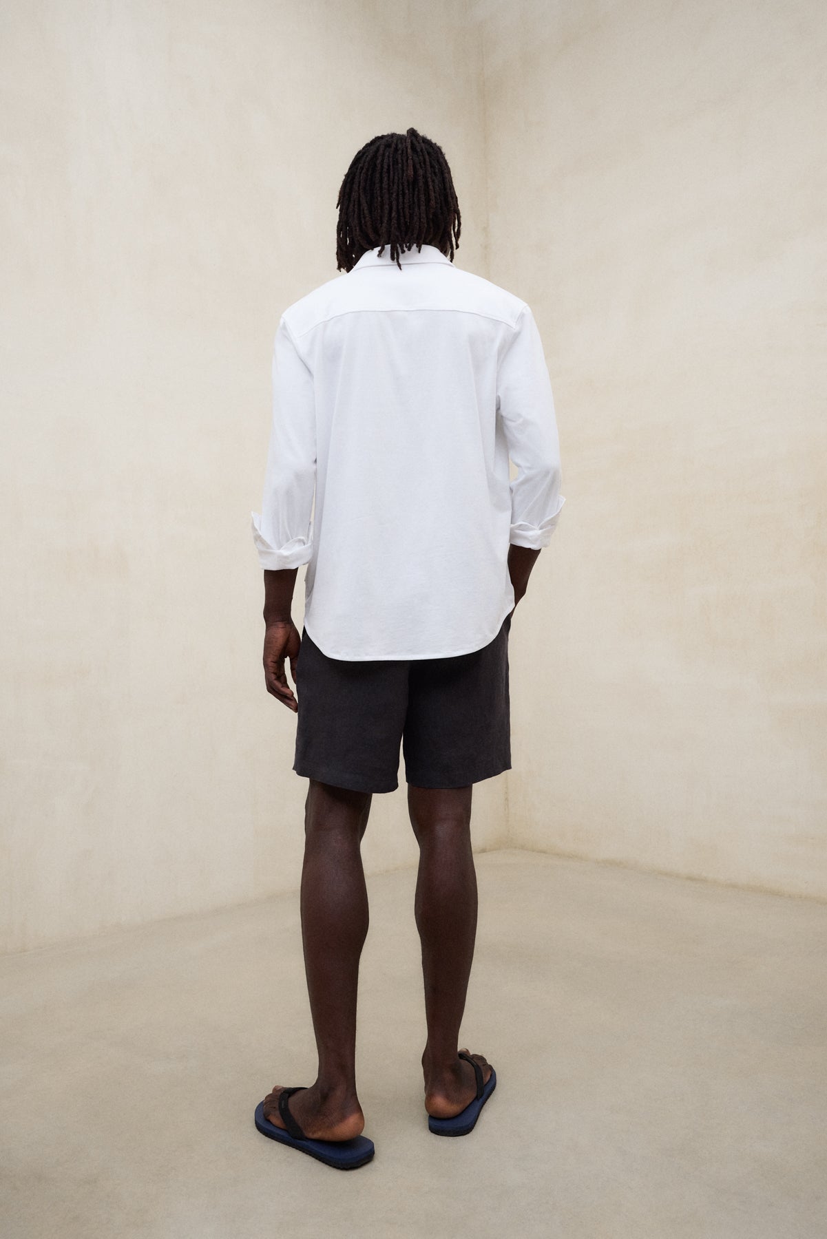 Un hombre de espaldas con camisa de Ecoalf blanca de manga larga, shorts oscuros y sandalias negras, en una sala de paredes claras.