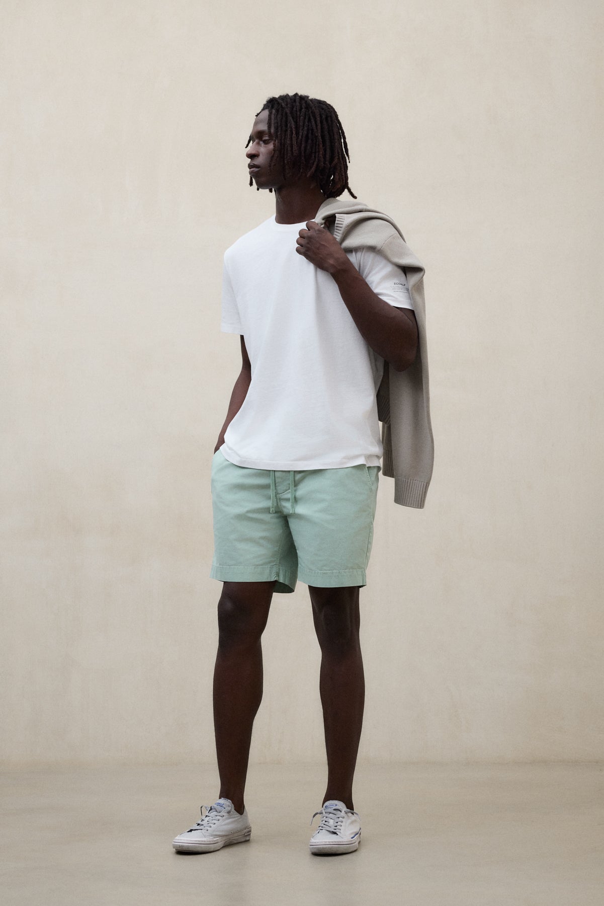 Un hombre con camiseta blanca y shorts verdes sostenibles de Ecoalf posando con un jersey gris en la mano sobre fondo neutro.