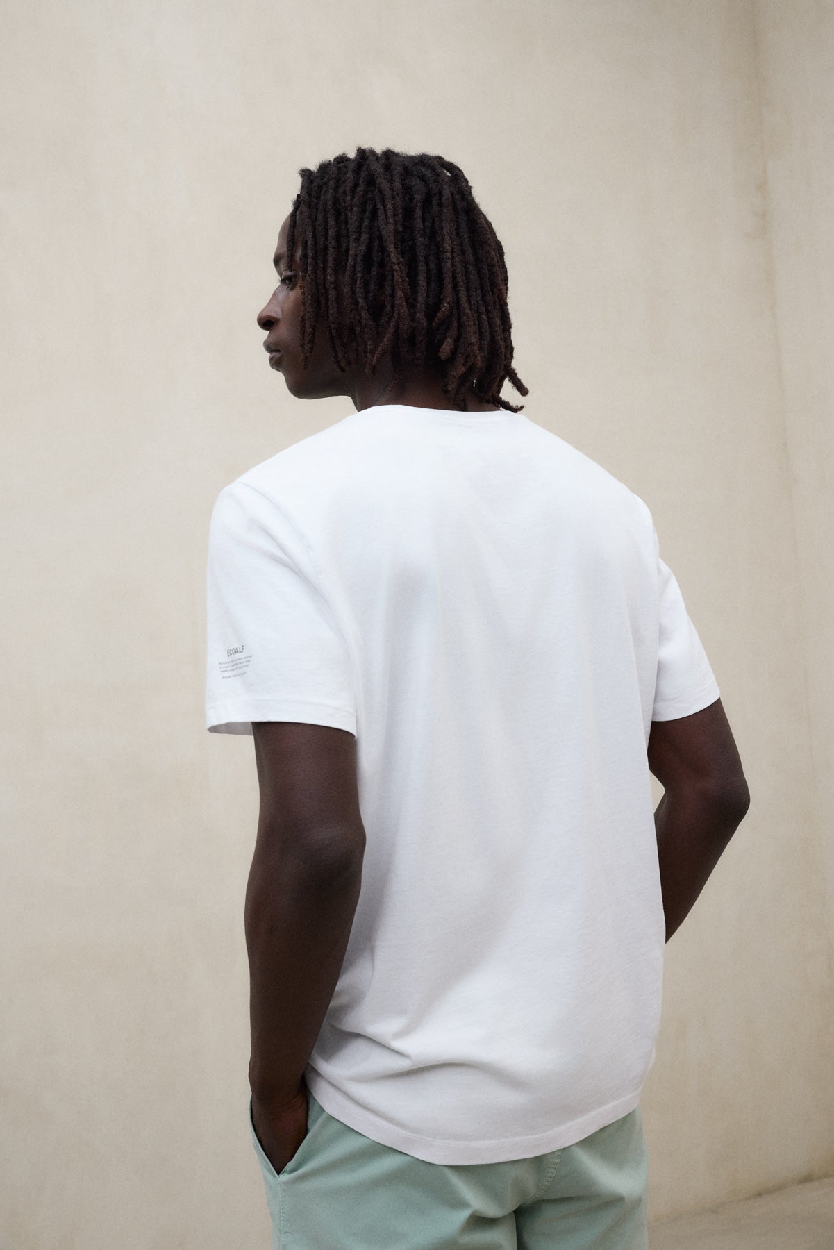 Un hombre de espaldas con camiseta blanca sostenible de Ecoalf y pantalón verde claro en un entorno minimalista.