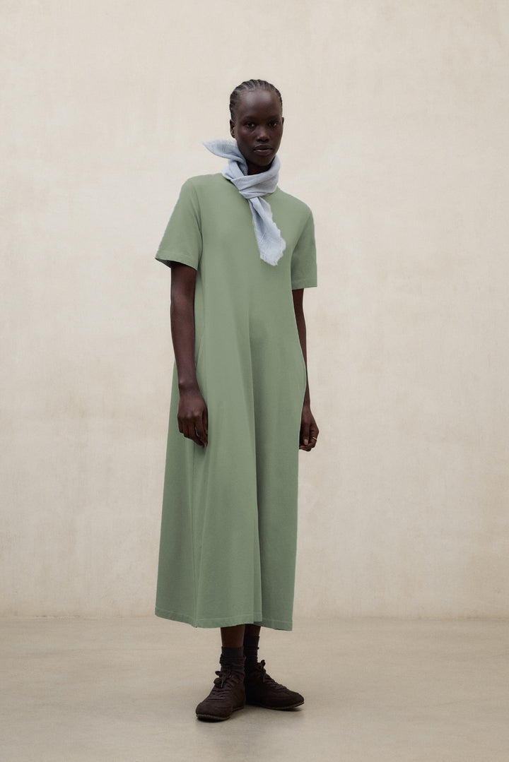 Una mujer en una sala beige con un vestido de Ecoalf largo verde oliva y un pañuelo ligero gris claro, con calcetines a juego y zapatillas oscuras.