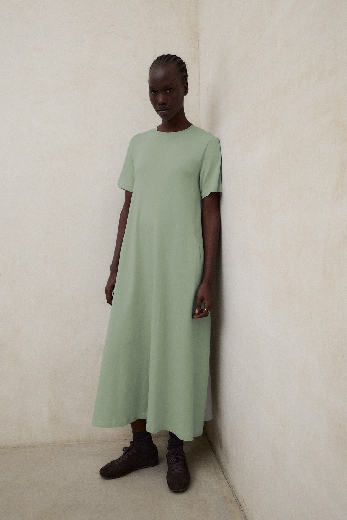 Una mujer en un rincón con un vestido de Ecoalf largo verde claro y zapatillas negras, ante una pared clara.