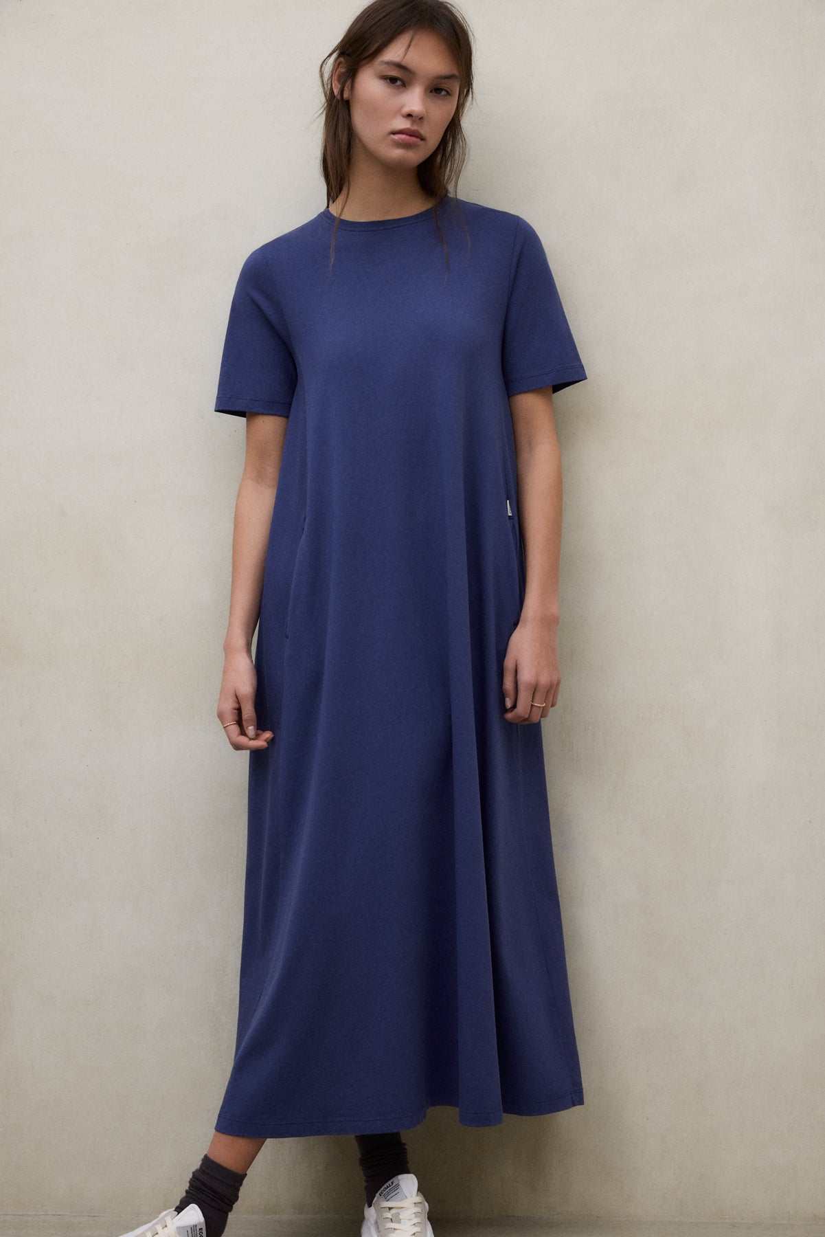 Una mujer con un vestido de Ecoalf largo azul de manga corta frente a una pared neutra, con calcetines oscuros y zapatillas blancas.