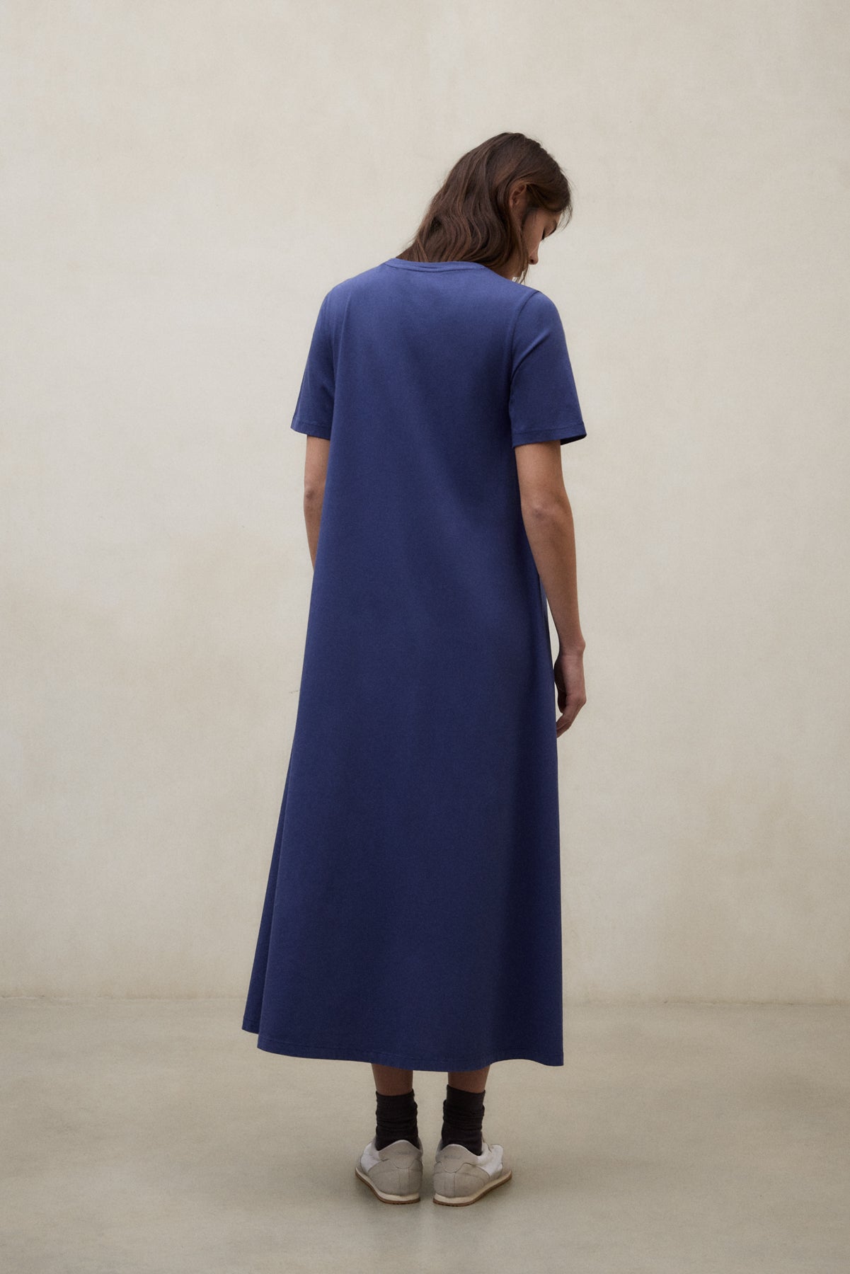 Una mujer de espaldas con un vestido de Ecoalf azul de manga corta hasta los tobillos, calcetines oscuros y zapatillas blancas, sobre fondo beige claro.