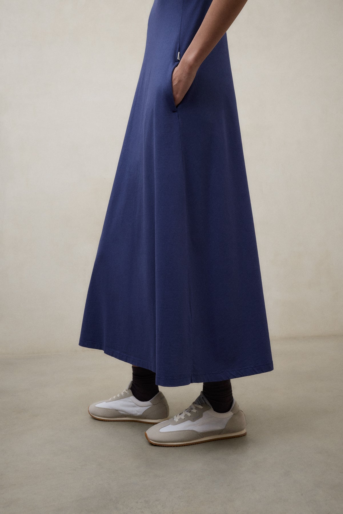 Una mujer con un vestido de Ecoalf largo azul con bolsillos, zapatillas blancas y calcetines oscuros, sobre un suelo claro.
