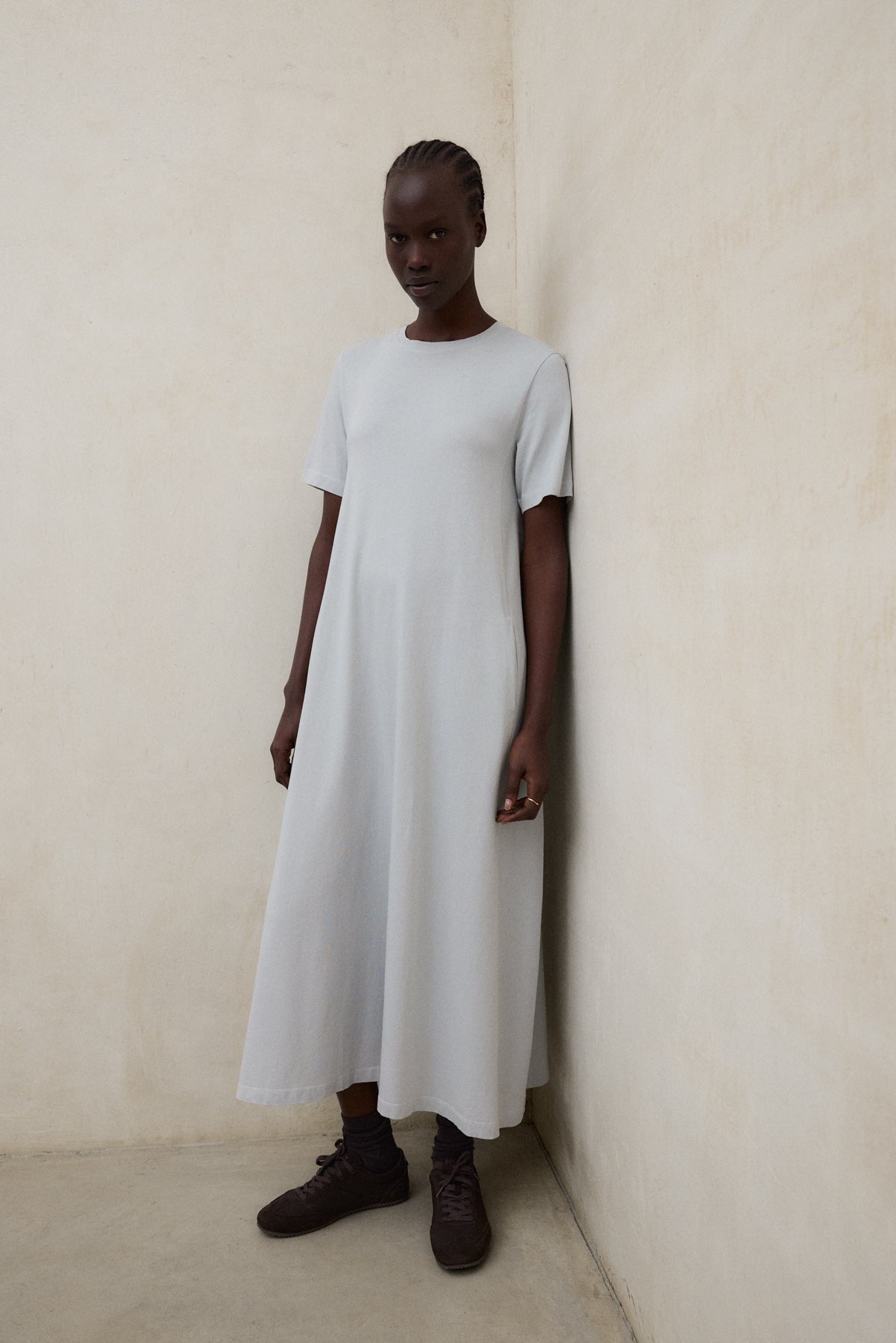 Una mujer frente a una pared clara con un vestido de Ecoalf largo gris claro, zapatillas oscuras y calcetines a juego, en un ambiente minimalista.