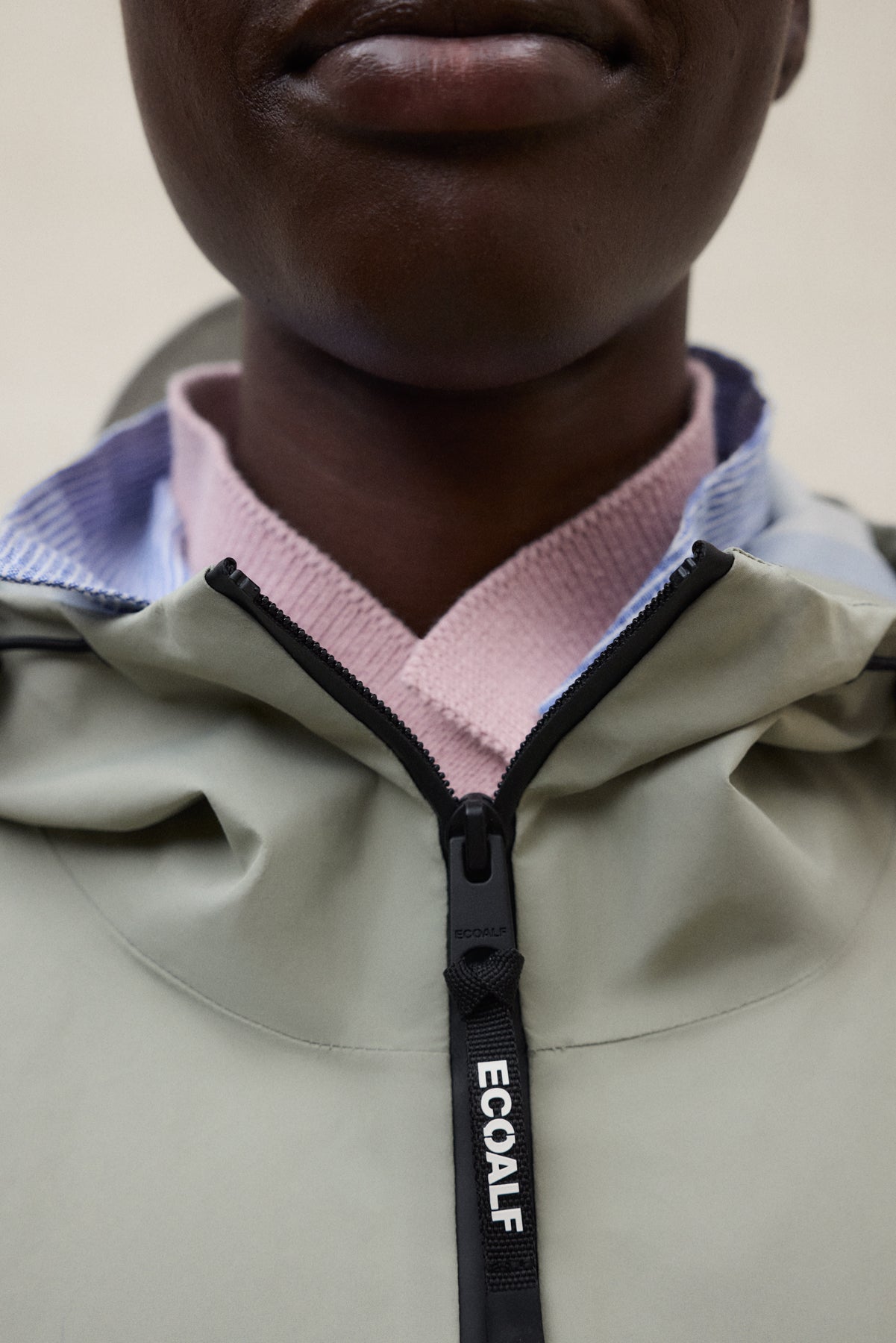 Primer plano de una chaqueta verde claro con la cremallera negra de la marca "ECOALF"; por debajo asoma una camisa de cuello rosa y azul.