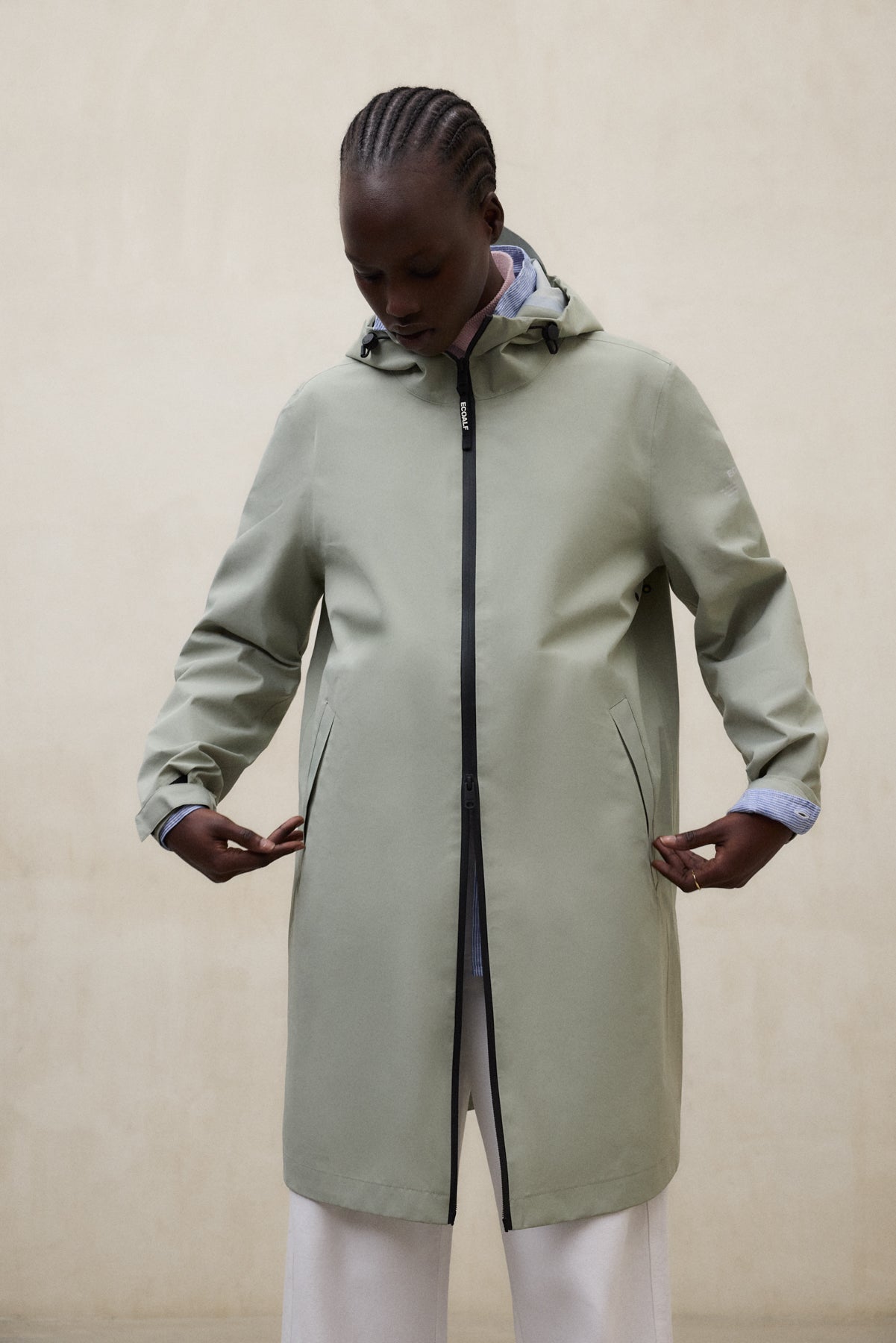 Una persona con un abrigo de Ecoalf largo verde claro con capucha y bolsillos con cierre, sobre una camisa azul y pantalón blanco, mira hacia abajo ante una pared beige.