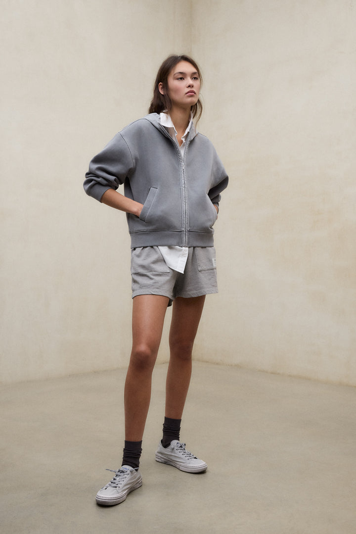 Un modelo con sudadera y shorts grises sostenibles de la marca Ecoalf, combinados con camisa blanca y zapatillas casual.