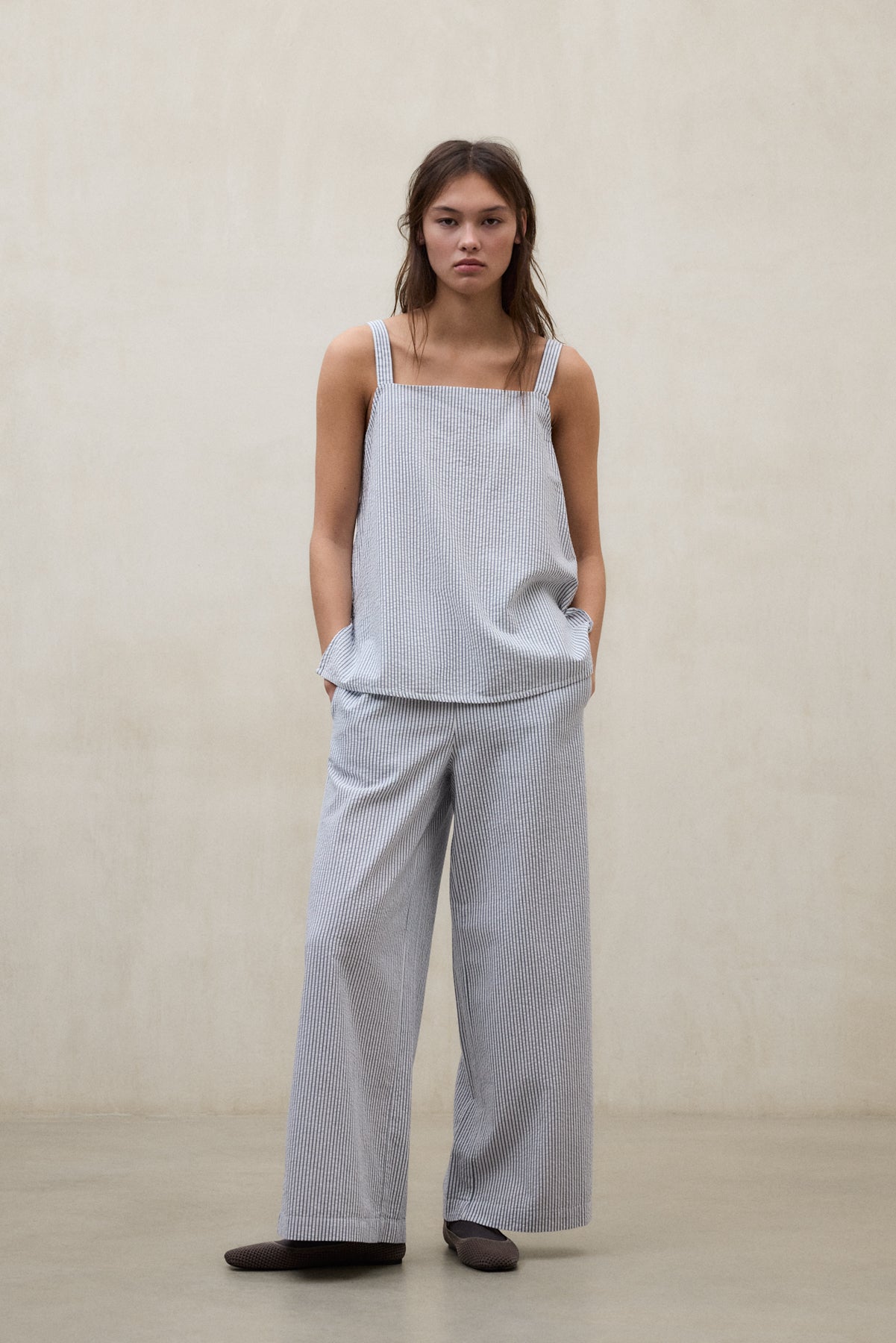 Una mujer con un conjunto de Ecoalf de pantalón y blusa a rayas azul claro y blanco, holgado, con calzado oscuro, en una sala de tonos neutros.