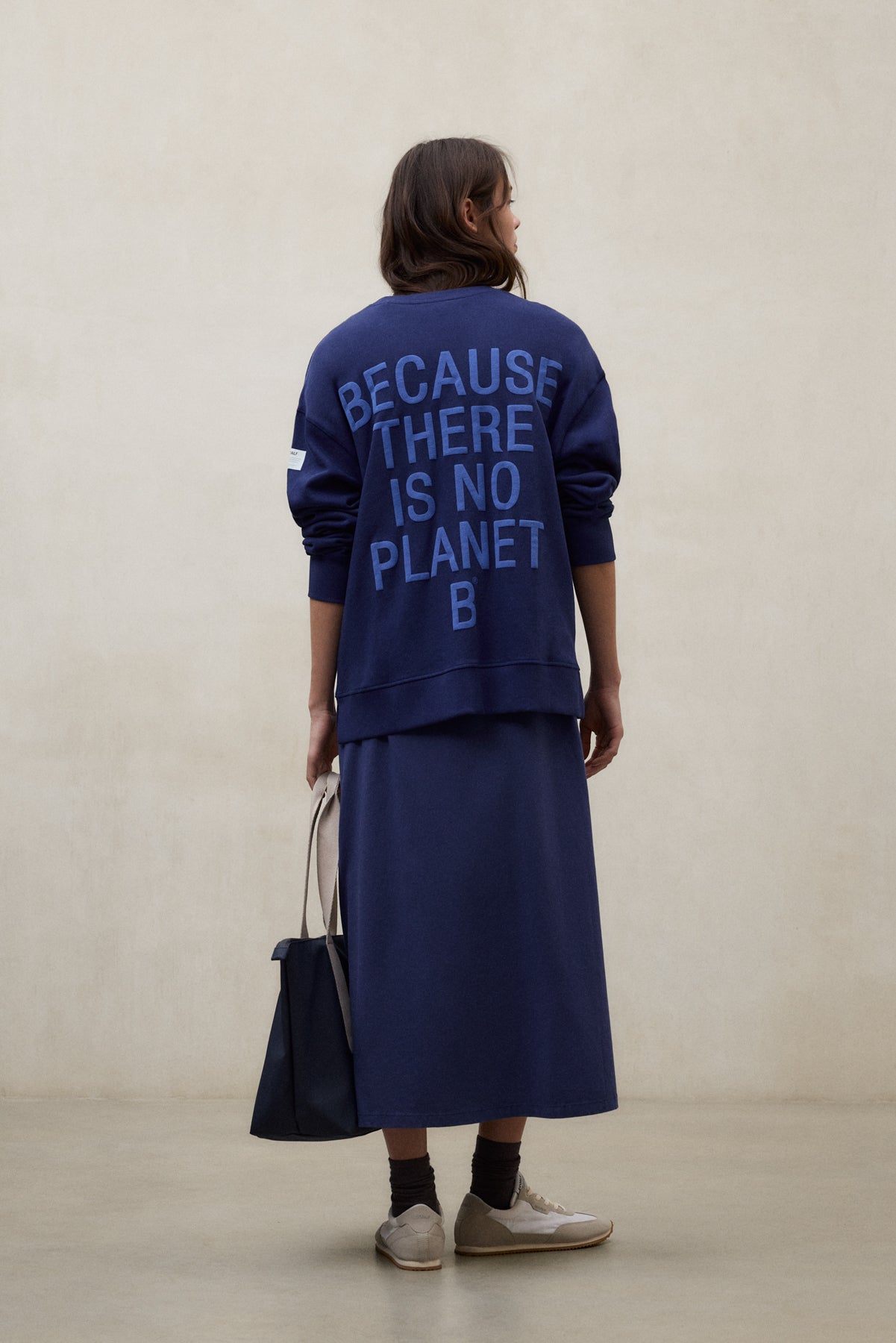 Una mujer de espaldas con sudadera azul de Ecoalf con el lema «Because There Is No Planet B», falda a juego y bolso en la mano.