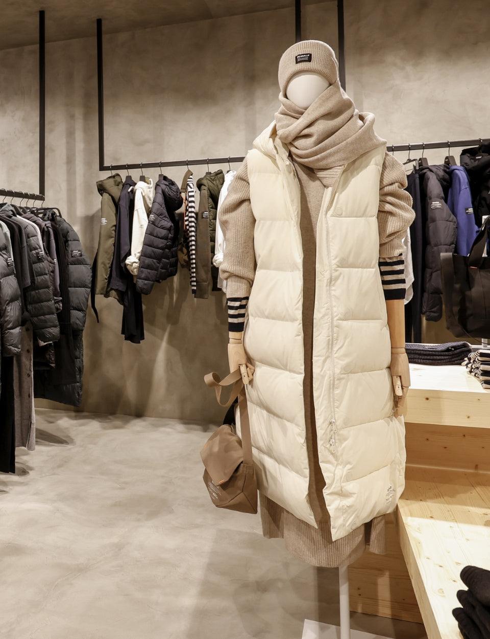 Un maniquí con un look de invierno —chaleco acolchado largo crema, jersey de punto beige y bufanda a juego— junto a chaquetas oscuras colgadas, en un espacio de cemento.
