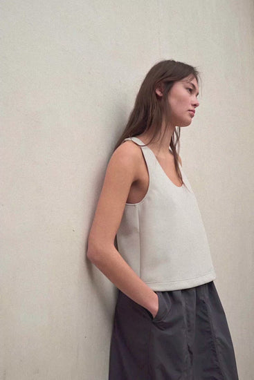 Una mujer de perfil apoyada en una pared, con una camiseta de tirantes de Ecoalf en tono claro y una falda oscura con bolsillos.