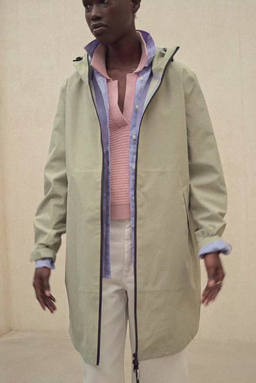 Un modelo con un abrigo de Ecoalf verde claro con capucha sobre una camisa azul y un jersey de punto rosa, con pantalón claro, sobre fondo neutro.