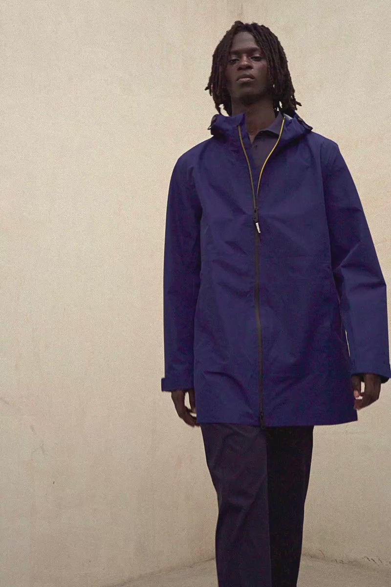 Un hombre con una chaqueta de Ecoalf azul oscuro con cremallera y pantalón a juego, frente a una pared clara.